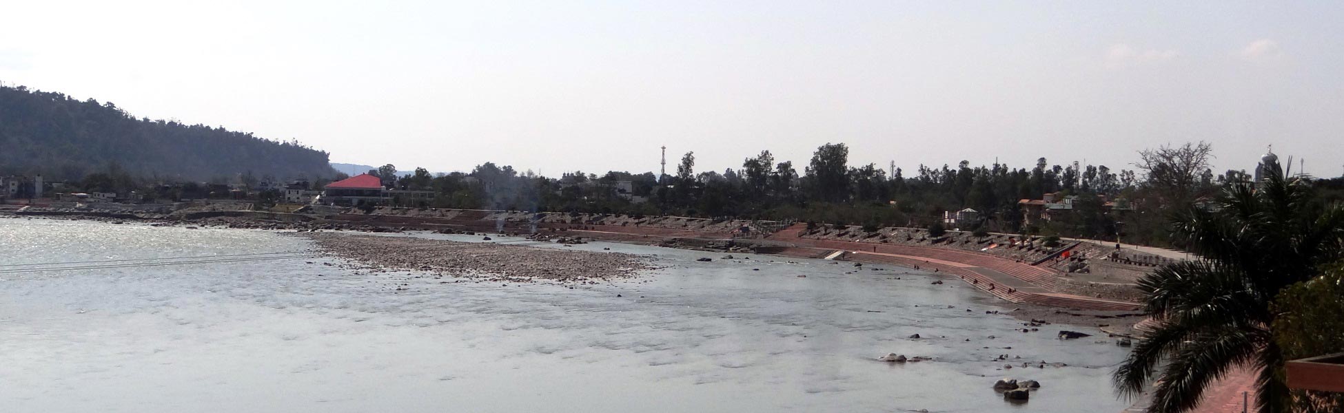 Wide panoramic view of a scenic riverfront film shooting location in India, featuring a sweeping curved concrete ghat embankment, a shallow rocky riverbed, and lush green hills with a red-roofed building in the background. film production house in Banka, video production house in Banka, tv production house in Banka, production house in Banka, film production company in Banka, video production company in Banka, tv production company in Banka, production company in Banka