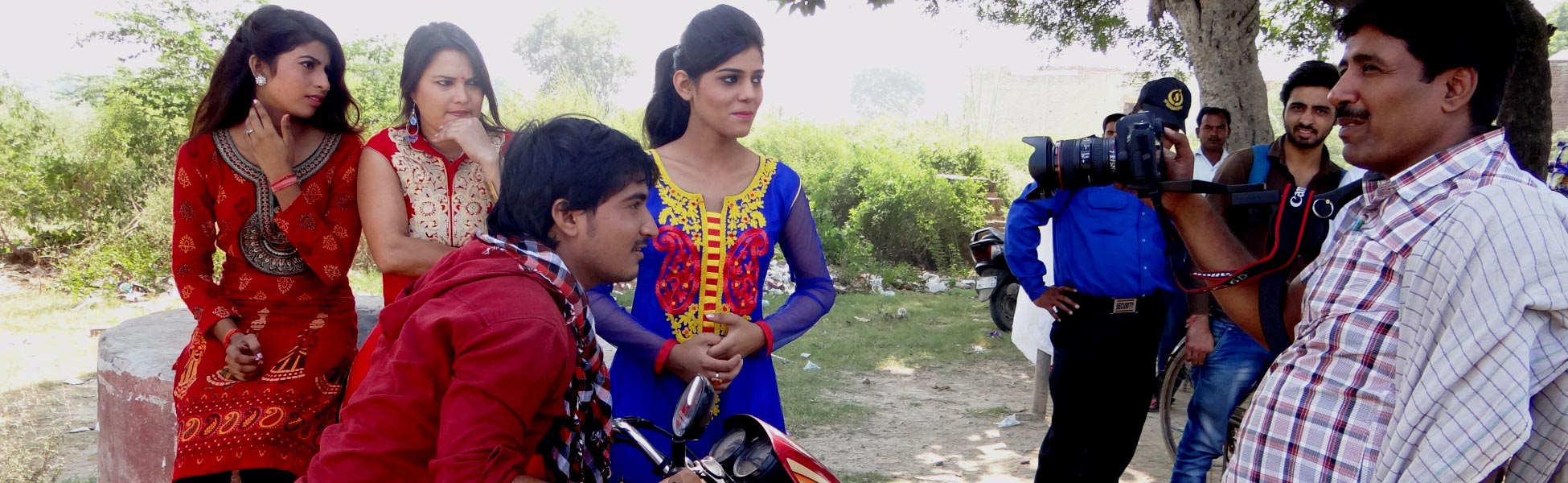 Behind-the-scenes view of an Indian short film crew filming a scene outdoors in Ghaziabad. A videographer uses a DSLR camera setup to film actors on a motorcycle and women in traditional attire. film shooting locations in Banka, video shooting locations in Banka, tv shooting locations in Banka, shooting locations in Banka