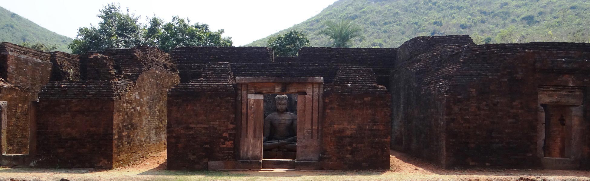 Ancient stone seated Buddha statue visible through a brick shrine doorway at the ruins of Monastery 1 in the Udayagiri Buddhist Complex, Jajpur, Odisha, India, with a lush green hill in the background. film production house in Bargarh, video production house in Bargarh, tv production house in Bargarh, production house in Bargarh, film production company in Bargarh, video production company in Bargarh, tv production company in Bargarh, production company in Bargarh