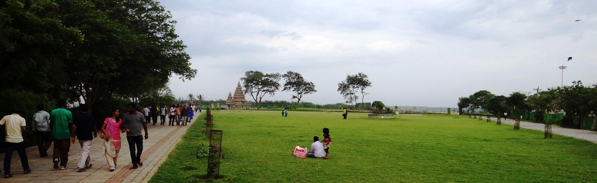 A wide-angle landscape view of the historic 8th-century Dravidian granite Shore Temple in Mahabalipuram, showing visitors on the paved pathway and an expansive green lawn under an overcast sky, ideal for large-scale cinematic set placement. film production house in Beed, video production house in Beed, tv production house in Beed, production house in Beed, film production company in Beed, video production company in Beed, tv production company in Beed, production company in Beed