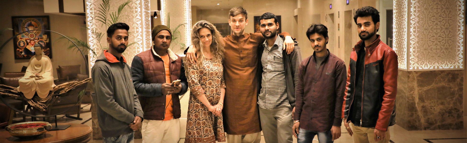 A group photo taken in a brightly lit hotel lobby features eight people: the professional camera crew from Trisna Films®, India, and their US client for a music video production in Bodhgaya, Bihar. They pose near a statue of a seated Buddha and decorative, illuminated pillars at the Lemon Tree Premier in Patna. film shooting locations in Champawat, video shooting locations in Champawat, tv shooting locations in Champawat, shooting locations in Champawat