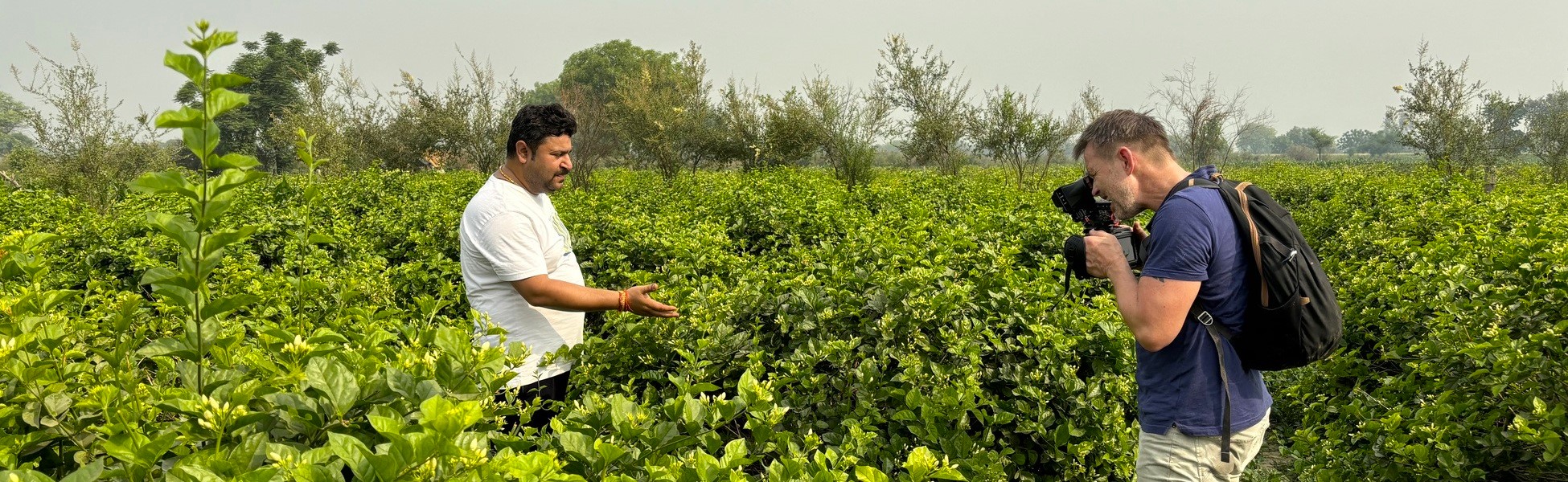 Director Mark Limburg holding a camera and filming a local man in a lush green field in Kannauj, Uttar Pradesh, India, during the production of the 'Rain' documentary with Trisna Films®.