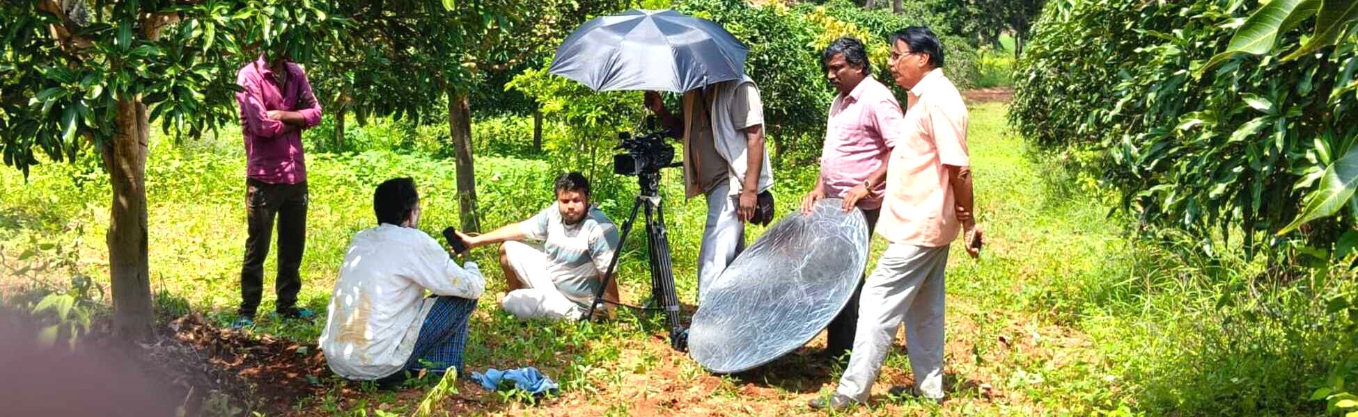 Trisna Films® crew providing on-location production support for an Austrian TV documentary on organic mango farming in Bengaluru South, Karnataka. The behind-the-scenes image shows the local Indian fixer team managing camera and lighting setups in a rural agricultural orchard. tv series shooting locations in india