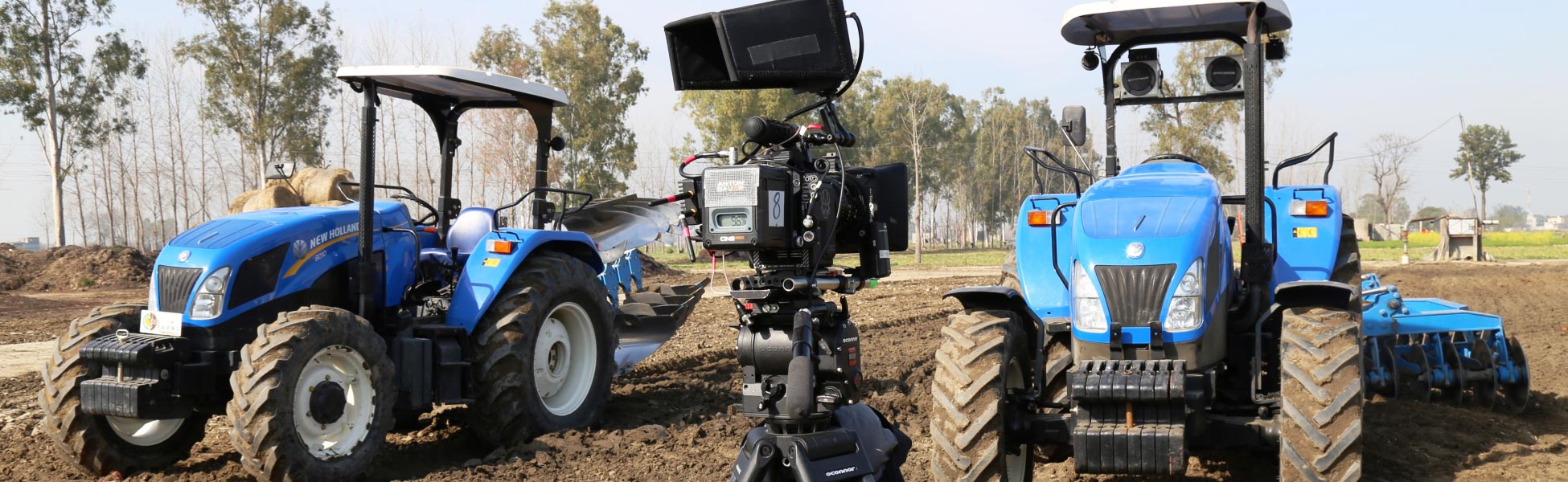 A professional cinema camera set up in a plowed agricultural field in Ludhiana, Punjab, flanked by two blue New Holland tractors during a web series shoot supported by Trisna Films®. film production house in australia, video production house in australia, tv production house in australia, production house in australia, film production company in australia, video production company in australia, tv production company in australia, production company in australia
