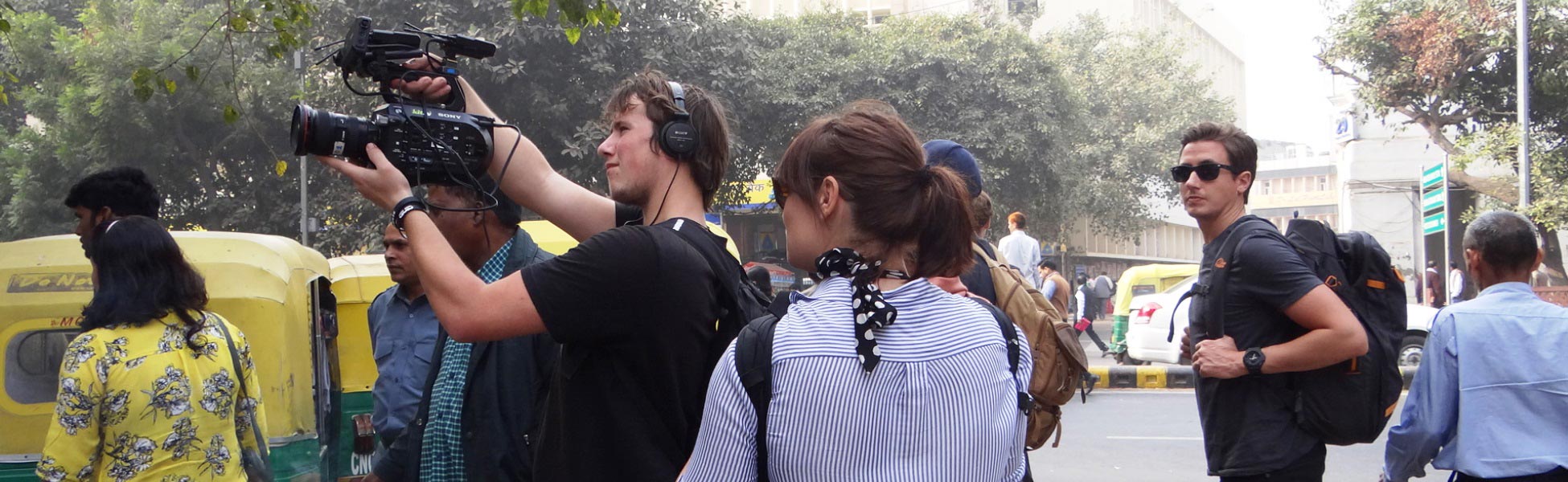 A British production crew from Insight TV filming on a bustling street in New Delhi, India. A cameraman holds a professional video camera, a crew member wears audio headphones, and iconic yellow-and-green auto-rickshaws are visible in the background. film shooting locations in Baghpat, video shooting locations in Baghpat, tv shooting locations in Baghpat, shooting locations in Baghpat