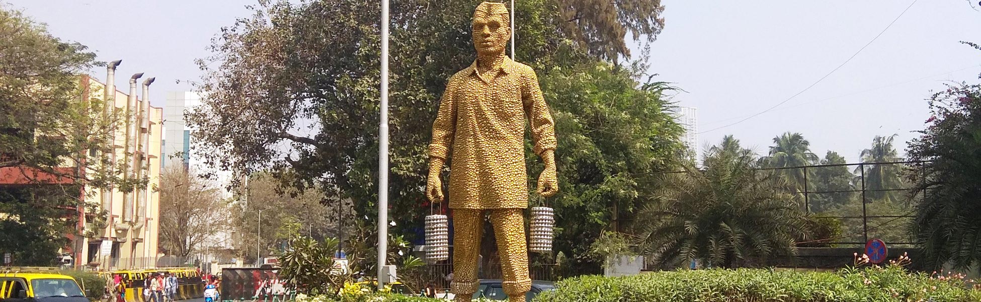 A wide daytime shot of the 13-foot-tall golden-colored stainless steel disc statue of a Mumbai Dabbawala wearing a traditional Gandhi topi and holding tiffin carriers, located at the Haji Ali junction roundabout on Tardeo Road in Mumbai, Maharashtra, India. film shooting locations in Balangir, video shooting locations in Balangir, tv shooting locations in Balangir, shooting locations in Balangir